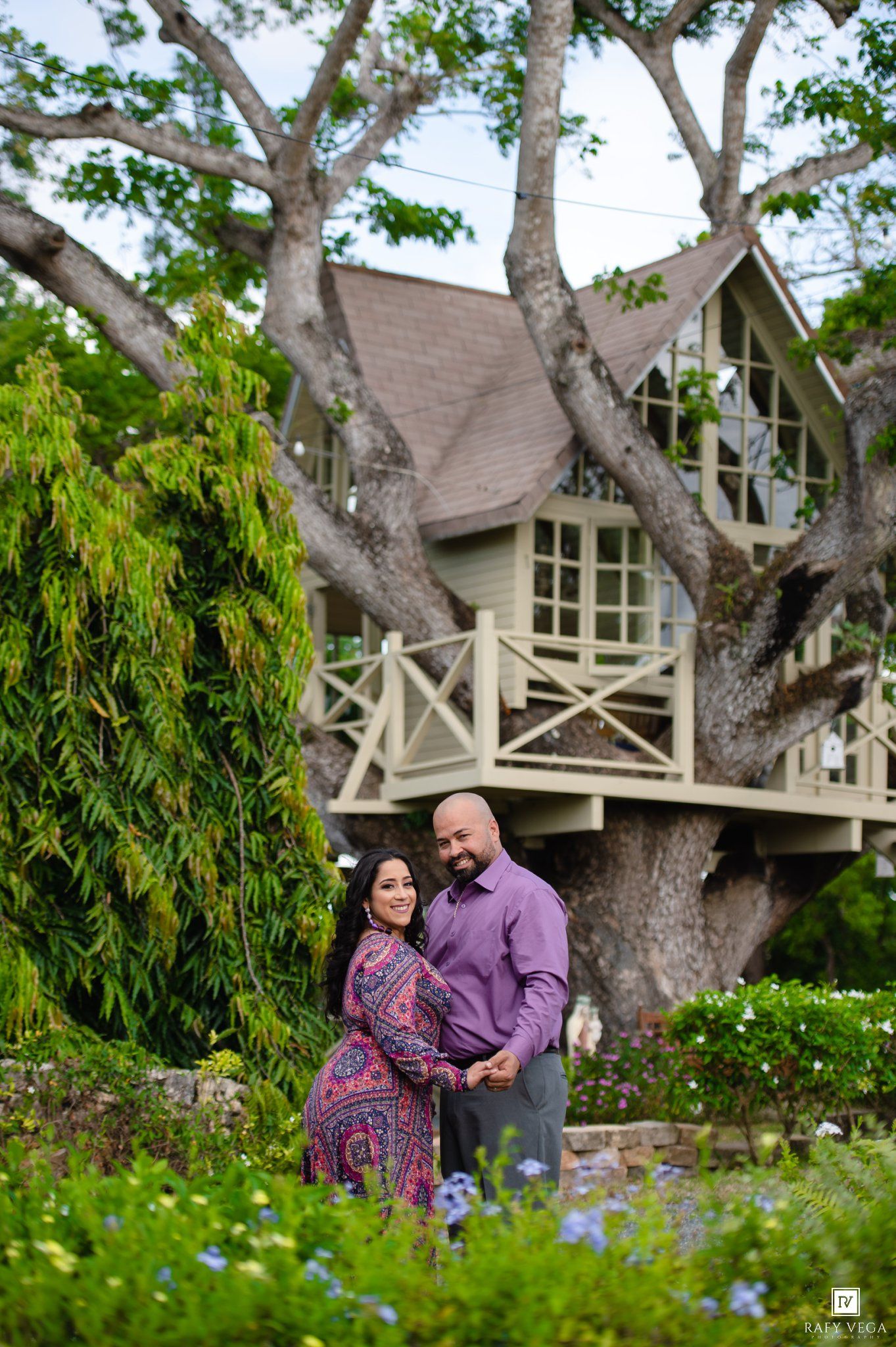 Couple poses romantically in lush garden before charming treehouse, woman in floral dress, man in purple shirt.