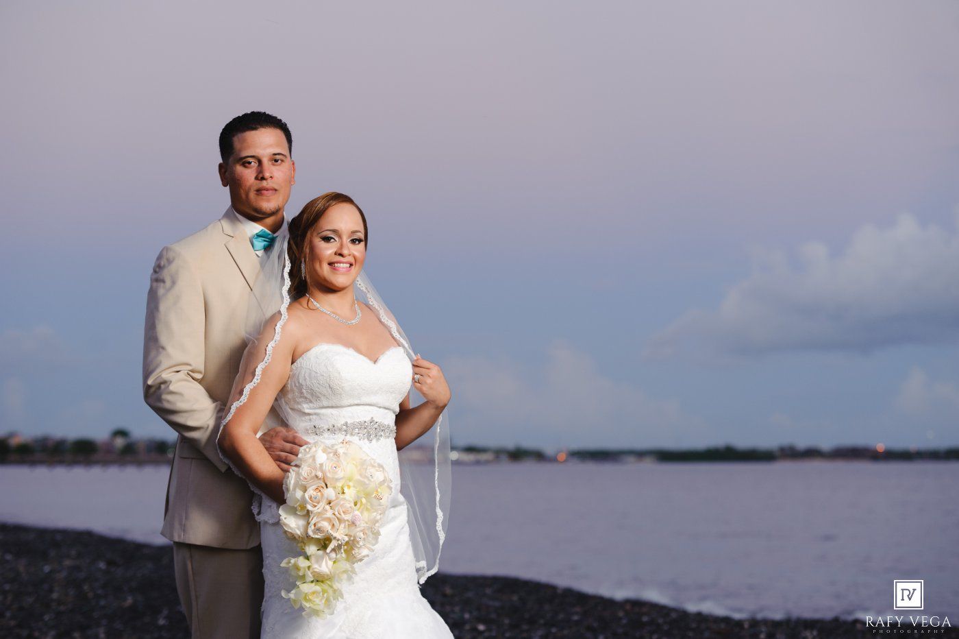 Bride and groom pose romantically on a beach at sunset, she holds a white bouquet in a strapless gown, he wears a tan suit.