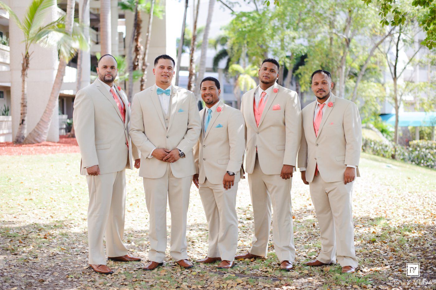 A groom and four groomsmen pose outdoors in matching beige suits with coral and turquoise accessories.