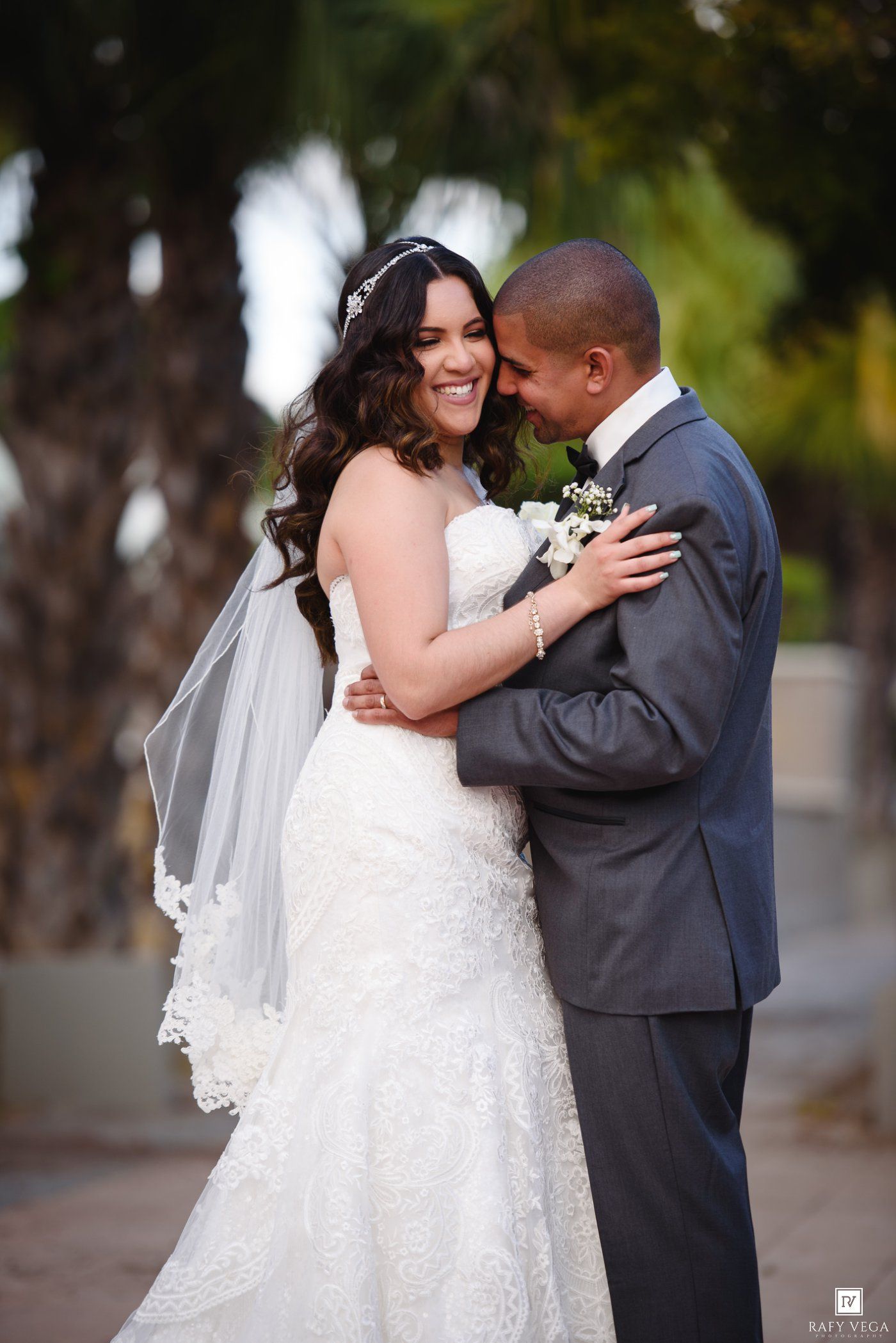 A smiling bride in a lace gown and veil embraces her groom in a gray suit outdoors at their wedding.