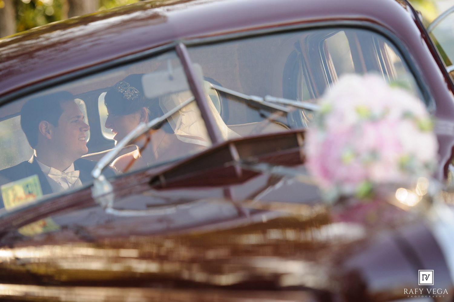 Couple inside a vintage brown car with a pink and white bridal bouquet visible in the foreground.