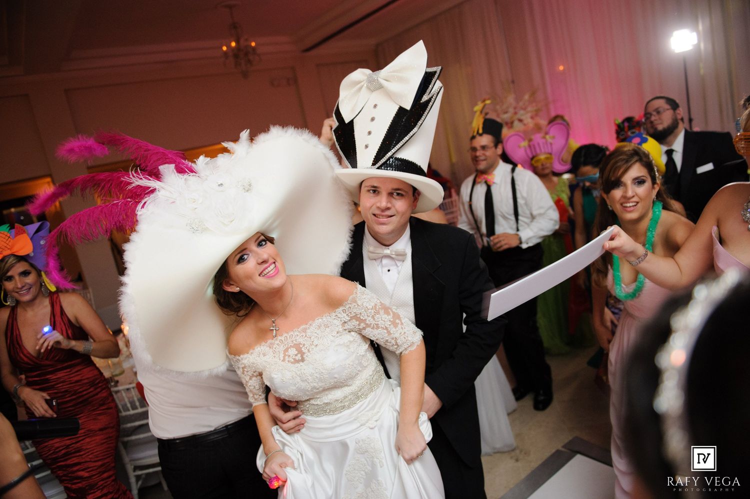 Bride and groom wearing festive oversized hats dancing at a colorful masquerade-themed wedding reception.