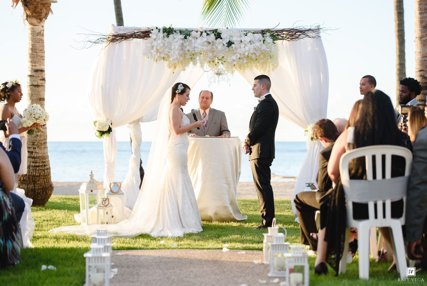 Elegant beach wedding ceremony under a white floral arch with ocean backdrop, bride and groom exchanging vows.