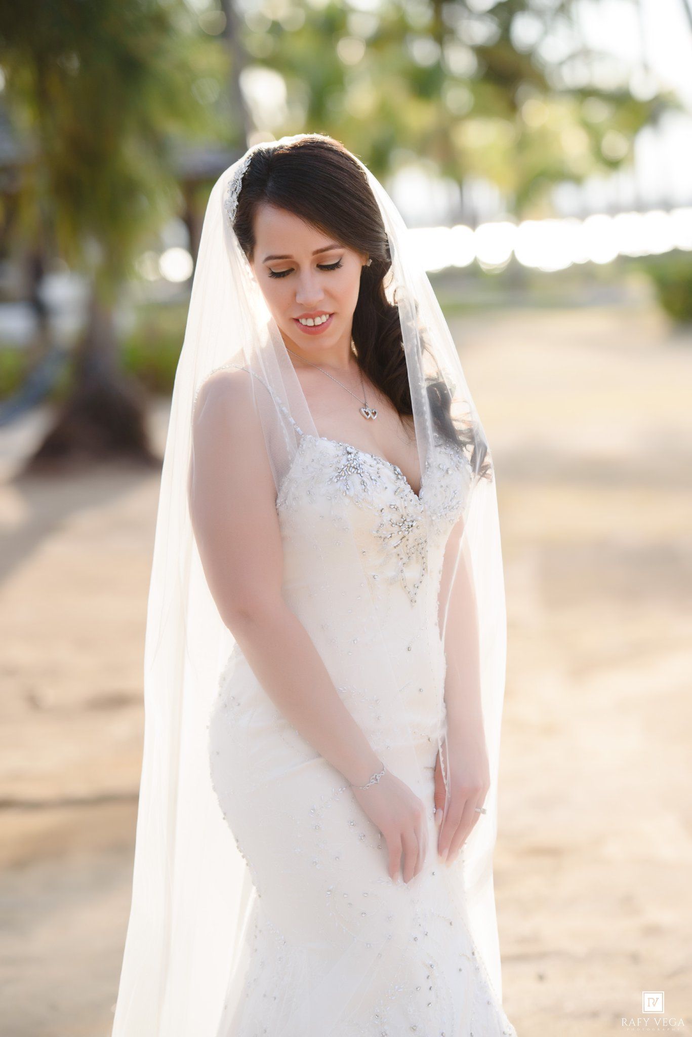 Bride in beaded mermaid gown and long veil poses on sandy beach with palm trees in golden light.