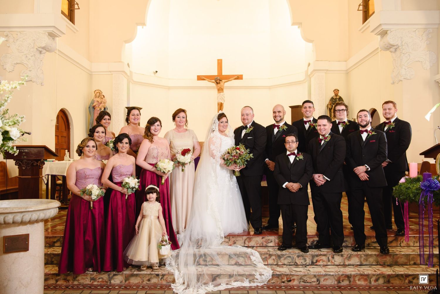 Wedding party poses in a church with bridesmaids in burgundy gowns, groomsmen in black tuxedos, and bride in white gown.