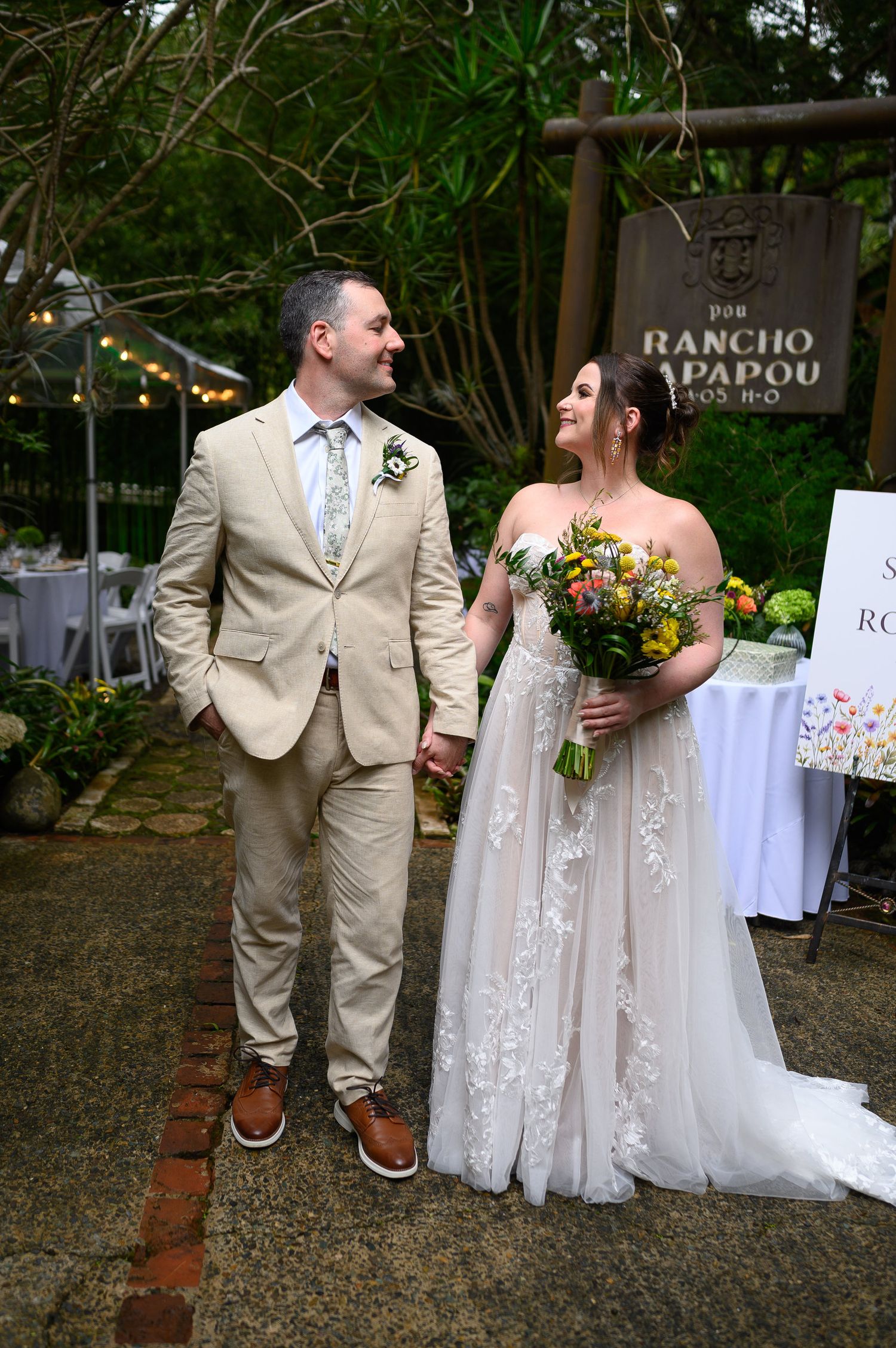 Bride in lace gown holding colorful bouquet stands with groom in tan suit at Rancho Papapou wedding venue.