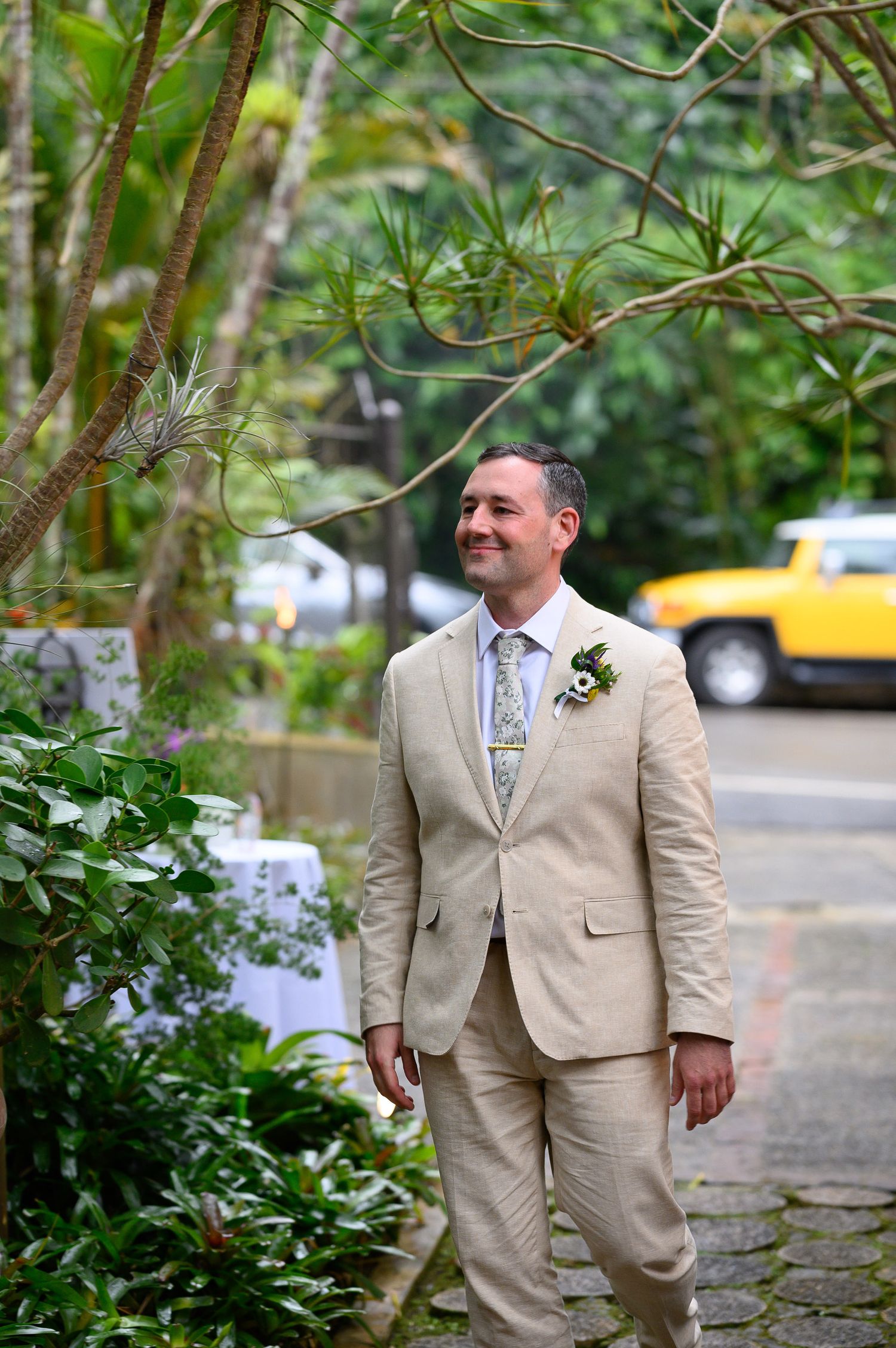 Groom in beige linen suit with floral boutonniere smiling in lush tropical garden setting.