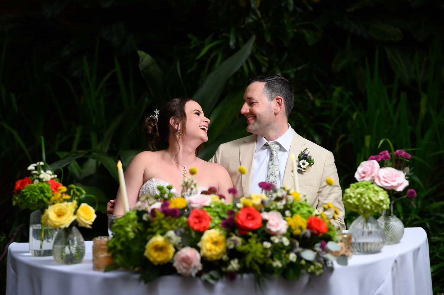 Bride and groom laughing together at floral sweetheart table with colorful wedding flowers outdoors.