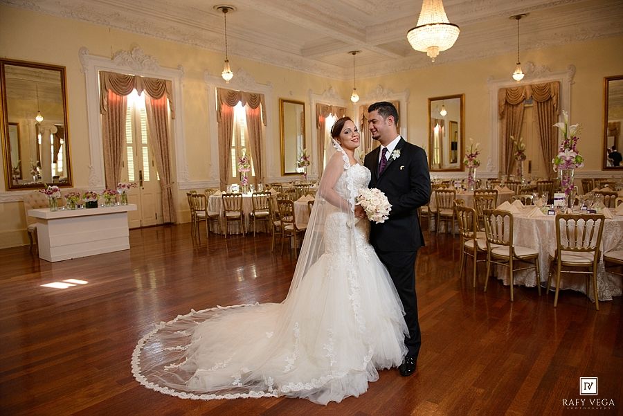 Bride and groom share romantic embrace on dance floor in elegant gold ballroom with chandeliers and draped windows.