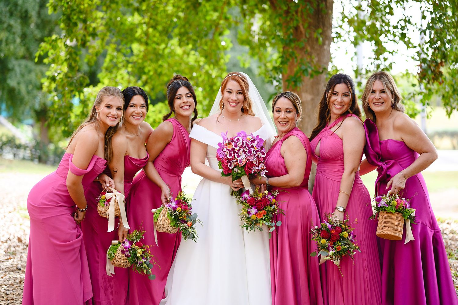 Bride in white gown poses with six bridesmaids in pink dresses holding colorful bouquets outdoors.