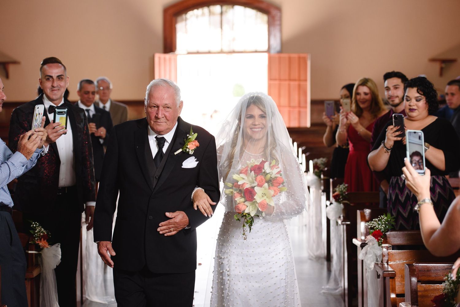 Bride in white lace gown walks down church aisle with older man, guests watching and photographing the ceremony.