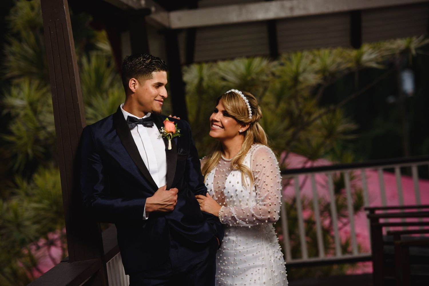 Bride in lace gown and groom in navy tuxedo share a romantic moment on an outdoor venue terrace at night.