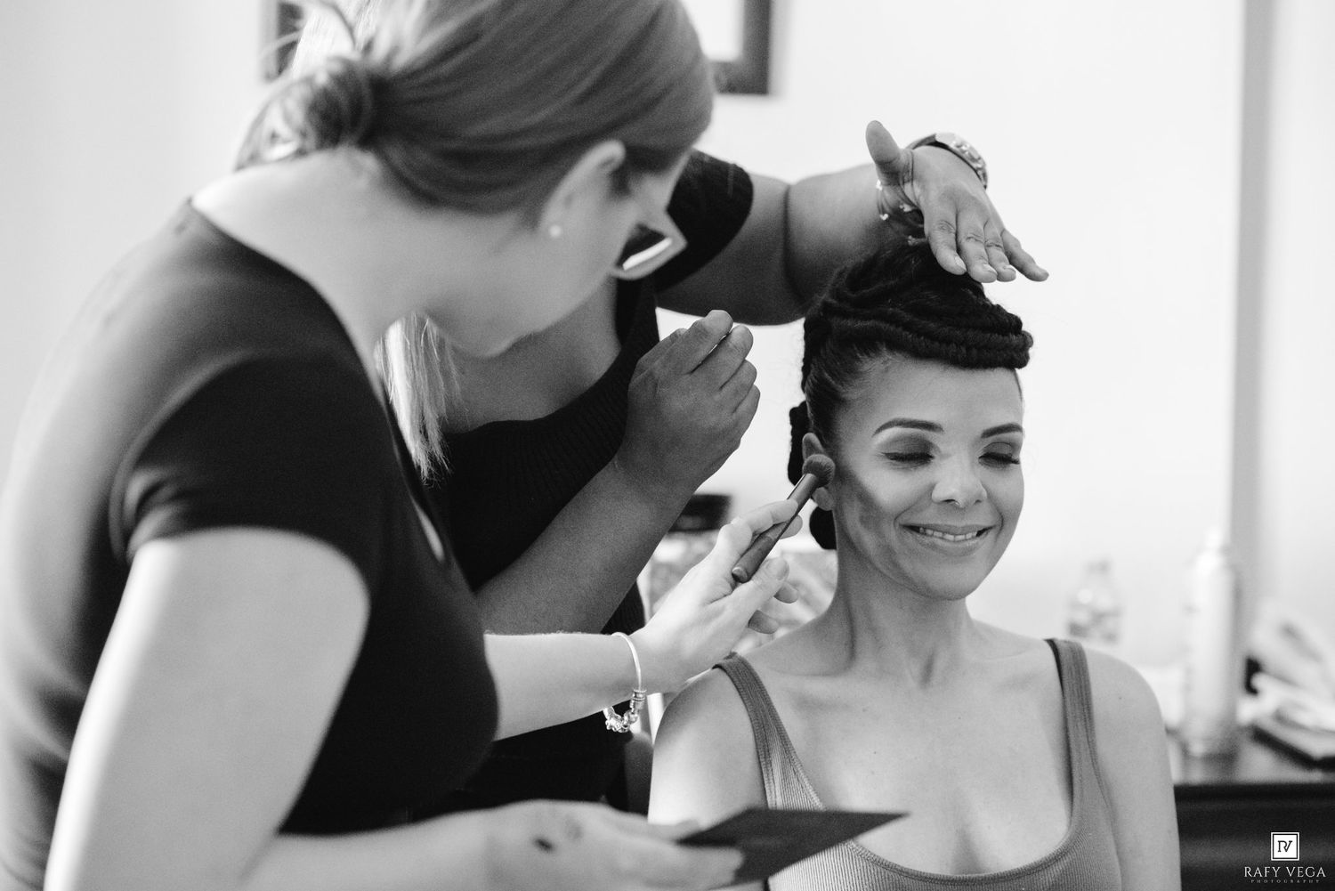 Black and white photo of a makeup artist applying makeup to a smiling bride during wedding preparations.