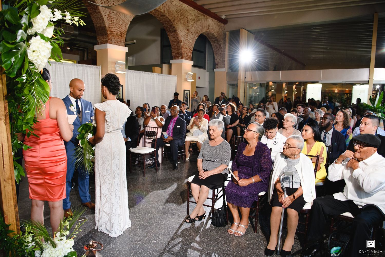 Elegant wedding ceremony with guests seated in rows, bride and groom at altar adorned with tropical floral arrangements indoors.