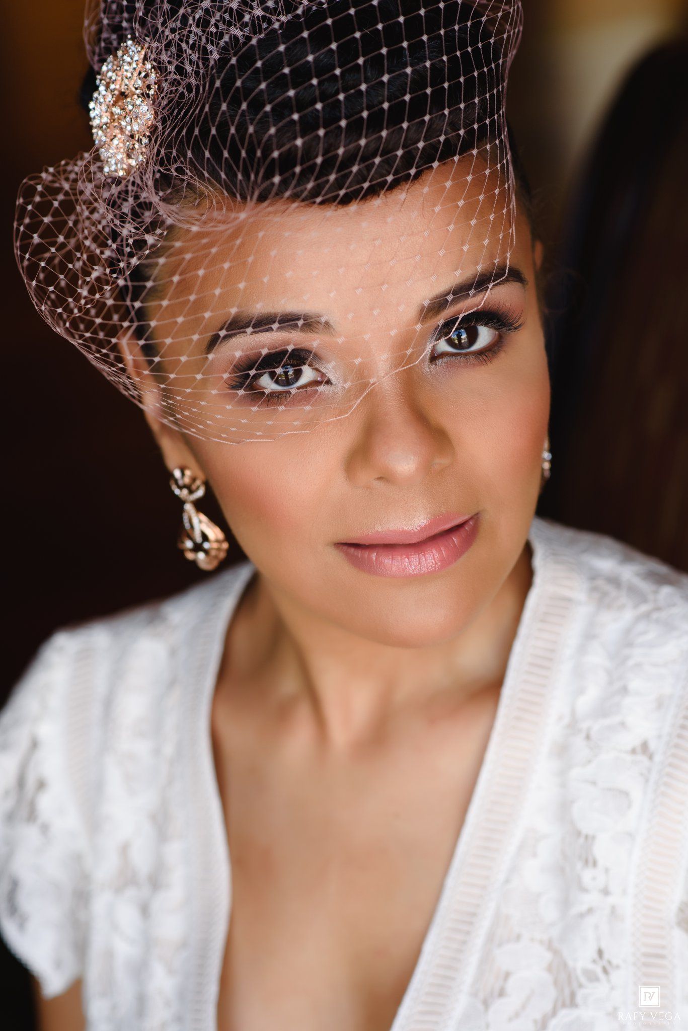 Elegant bride wearing a birdcage veil with jeweled accent and drop earrings, showcasing smoky eye makeup and lace dress.