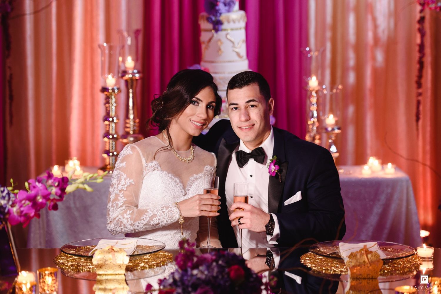 Newlyweds toast with champagne at candlelit wedding reception table with ornate cake and pink floral decor.