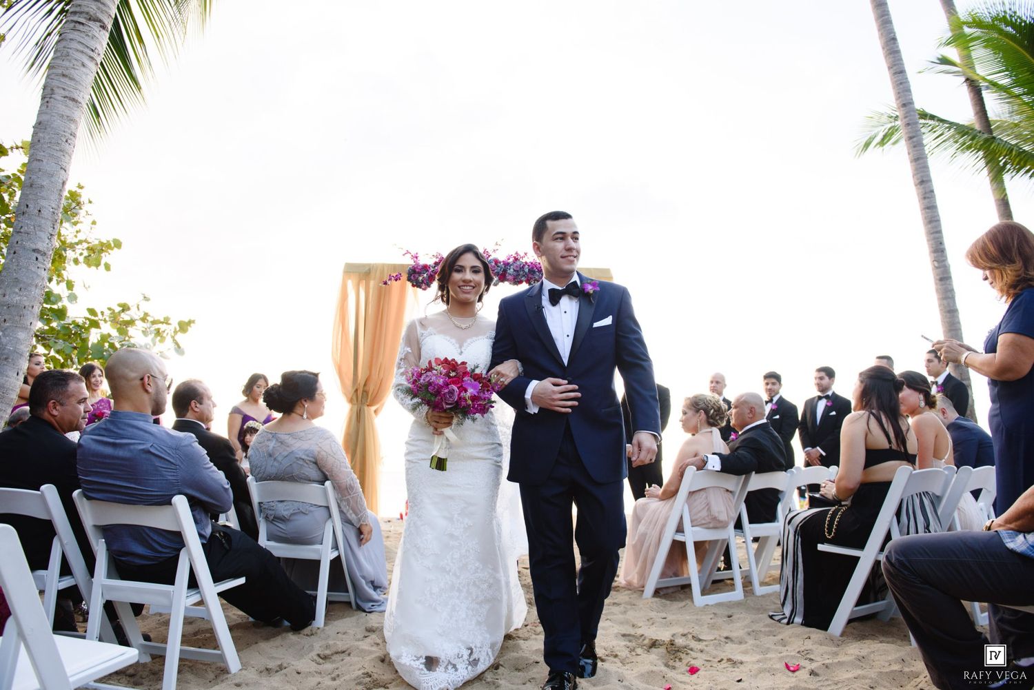 Bride and groom walk down sandy beach aisle after outdoor wedding ceremony with guests seated in white chairs.