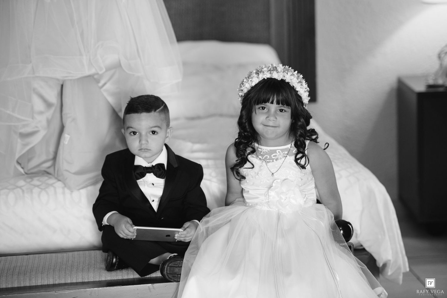 Black and white photo of a young boy in a tuxedo and girl in a white flower girl dress sitting together at a wedding.