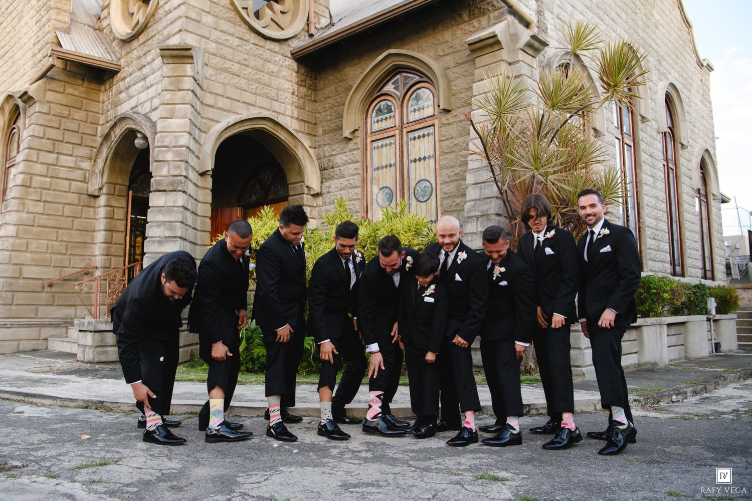 Groomsmen in black suits showing off colorful socks outside a stone church before a wedding ceremony.