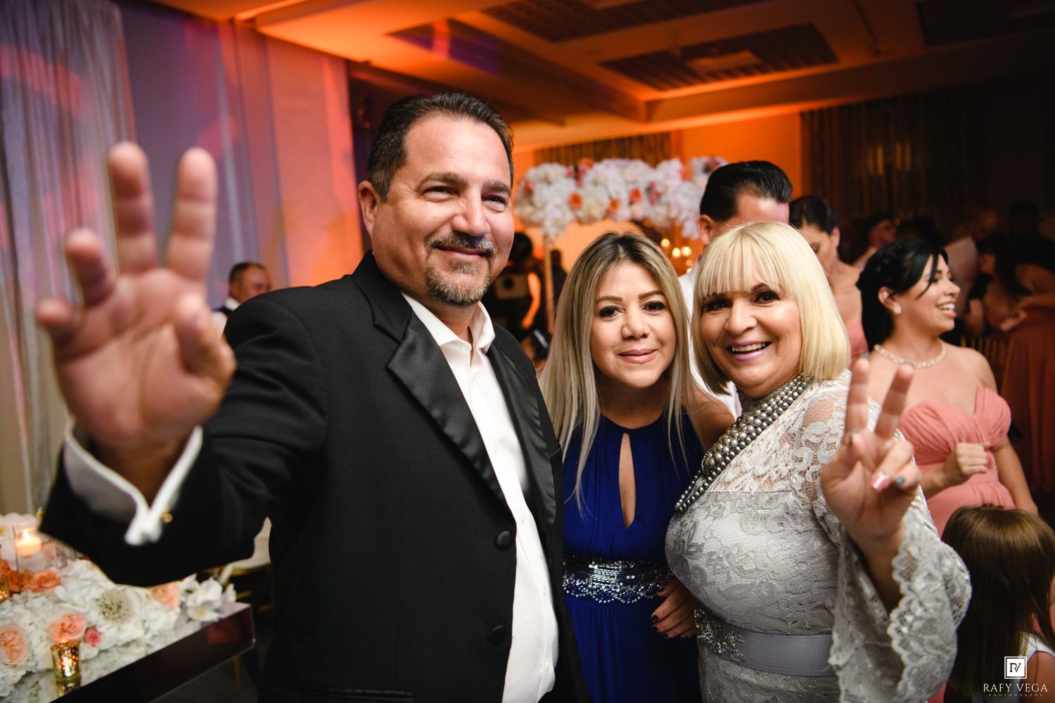 Three people smiling and posing with peace signs at an elegant indoor celebration event.