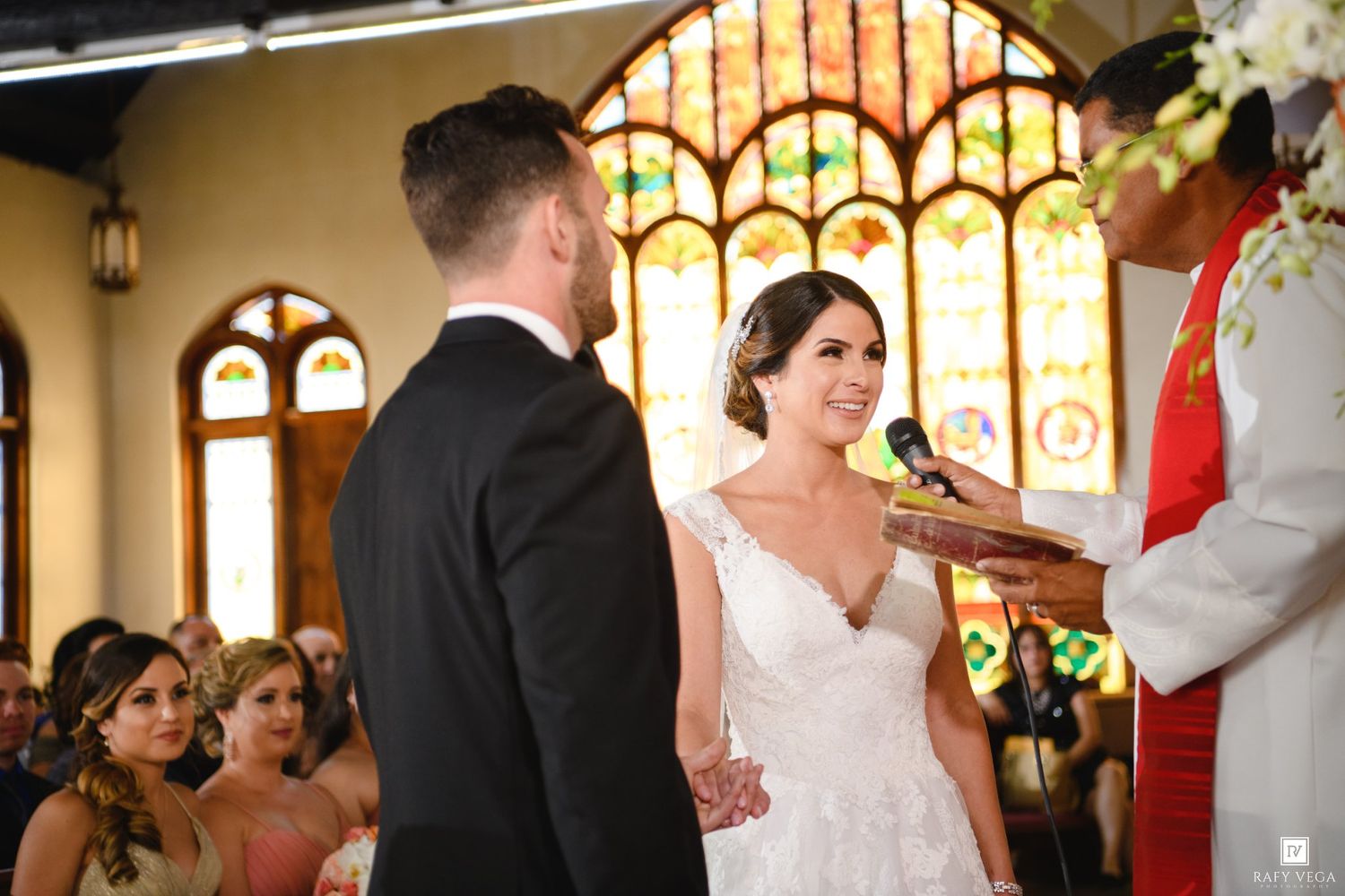Bride and groom exchange vows at church altar with colorful stained glass windows during wedding ceremony.