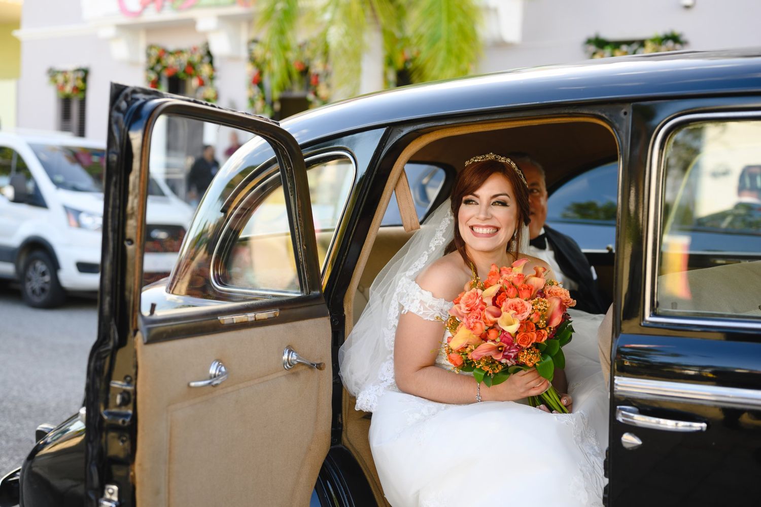 Smiling bride in white gown holding orange bouquet seated in classic black vintage car on wedding day.