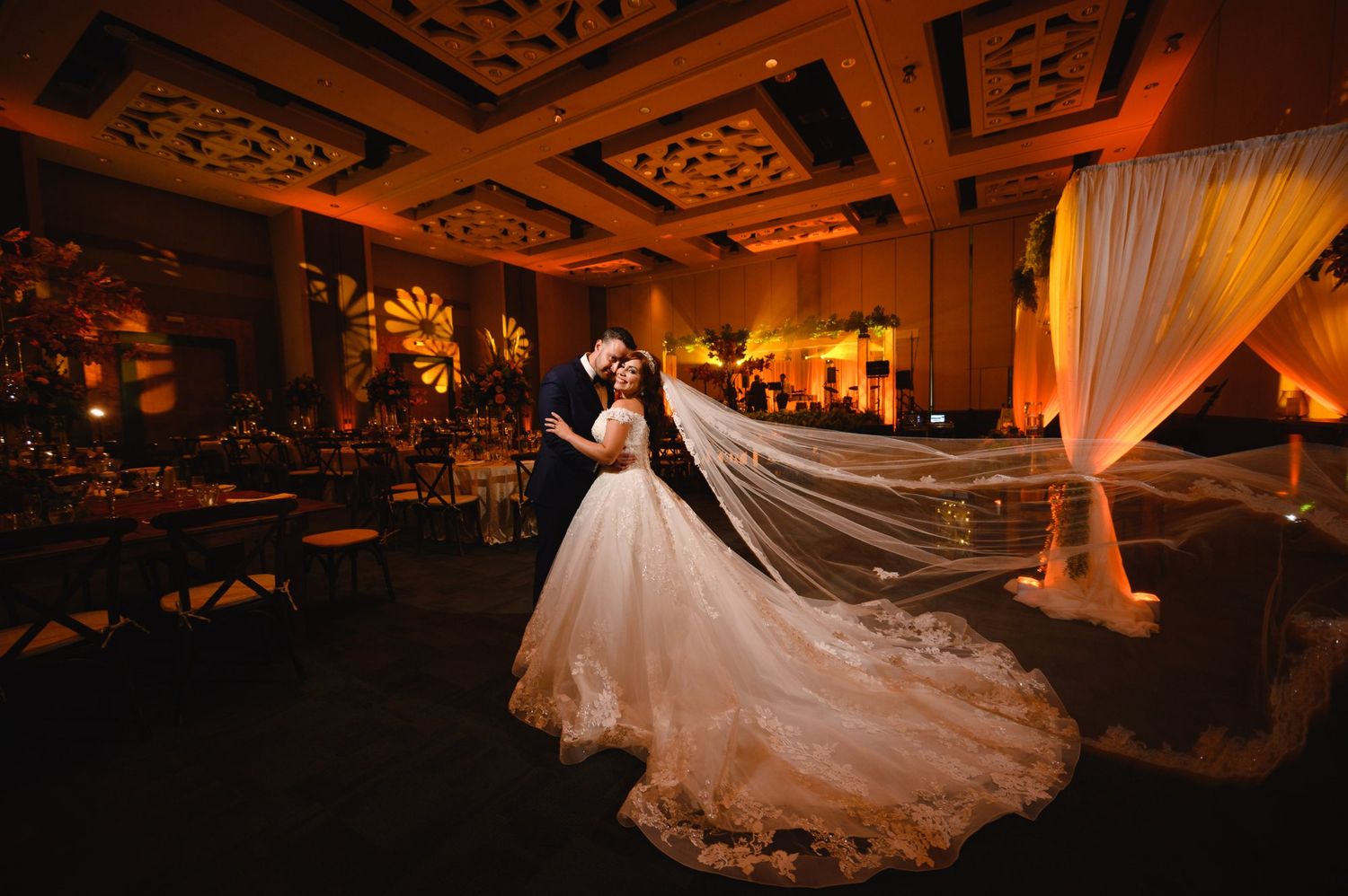 Bride and groom share romantic first dance in elegant ballroom with warm amber uplighting and flowing veil.