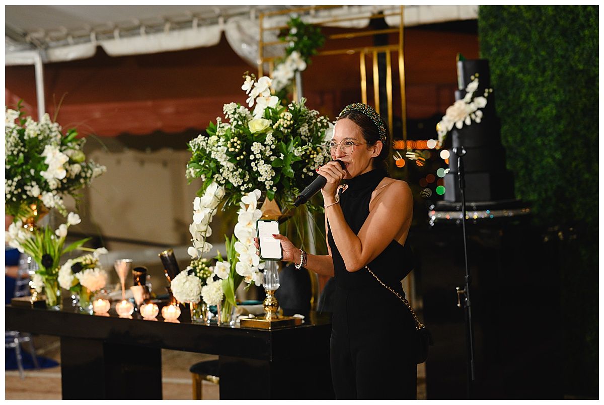 Woman in black dress speaking at microphone beside lush white floral arrangements at elegant evening event.