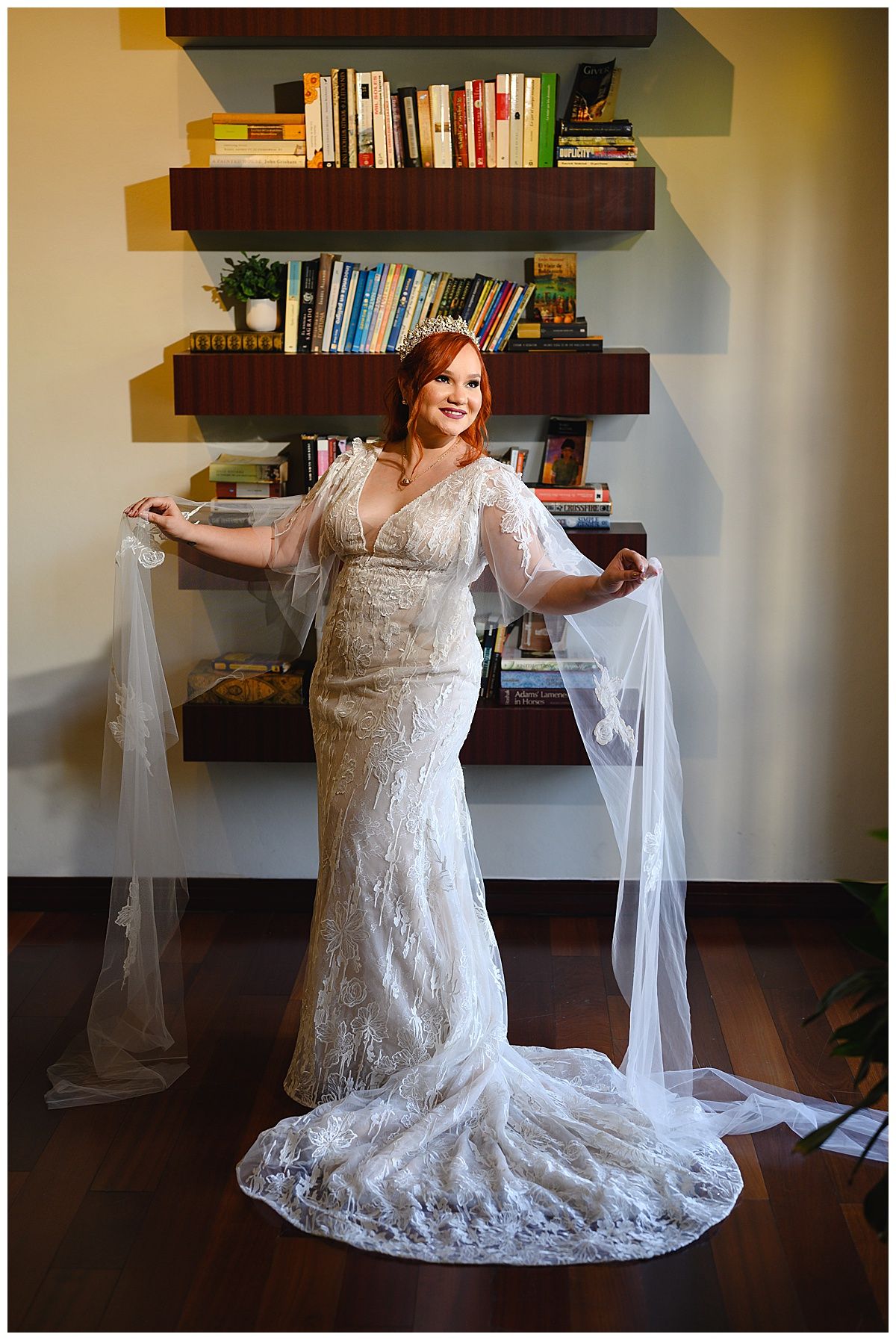 Bride in lace wedding gown with veil spread wide, standing before wooden bookshelves in elegant indoor setting.