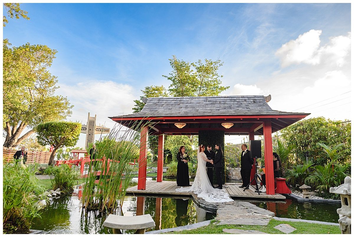 Wedding ceremony in a Japanese garden gazebo with red pillars, koi pond, and lush greenery under a blue sky.