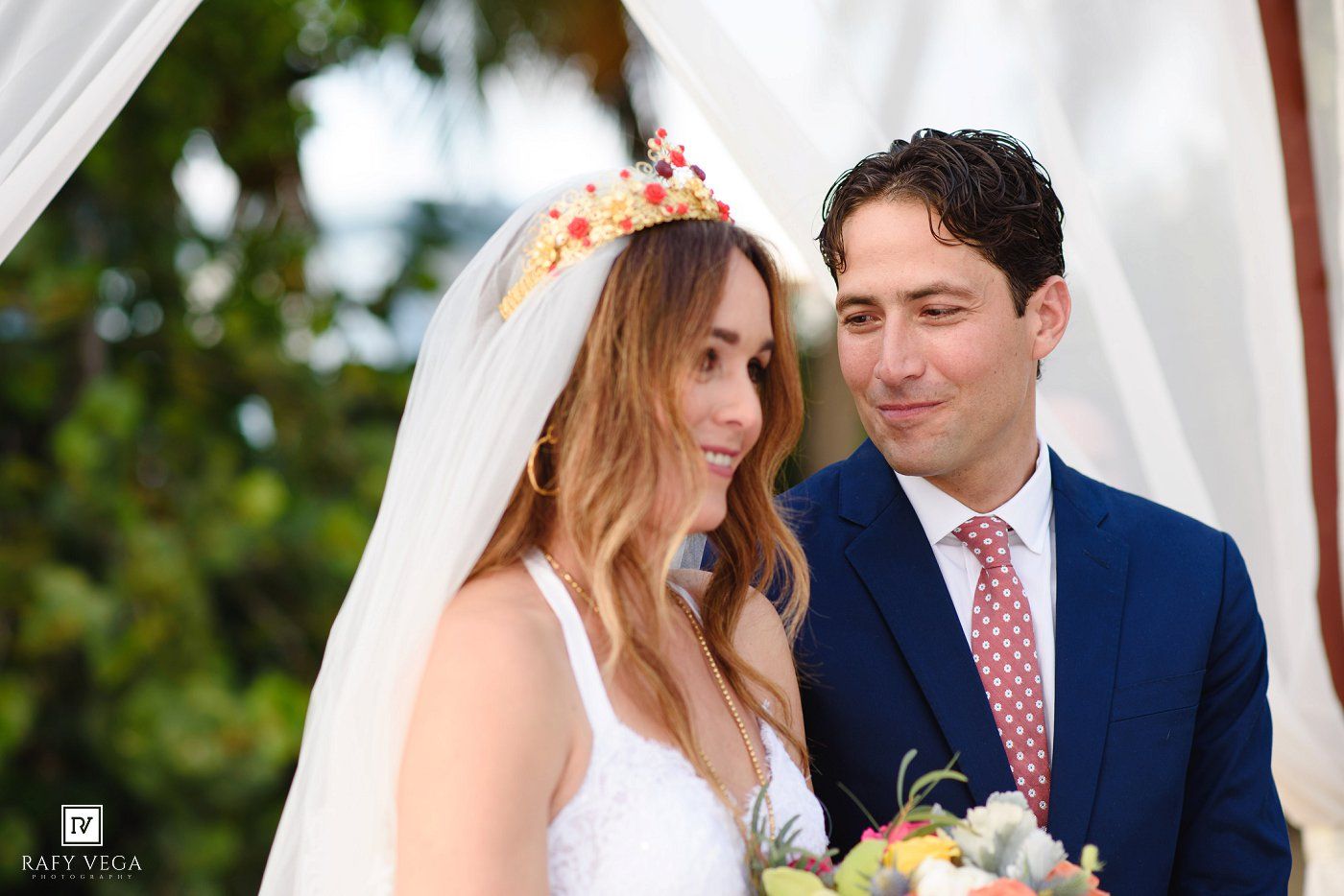 A bride in a floral crown and veil stands beside a groom in a blue suit at an outdoor wedding ceremony.