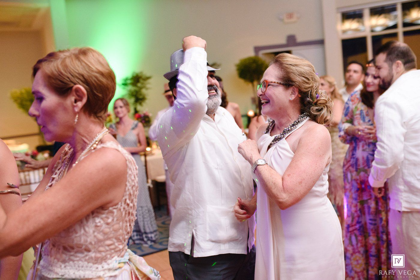 Guests dancing and celebrating joyfully at an elegant wedding reception with green lighting and decorated tables in the background.