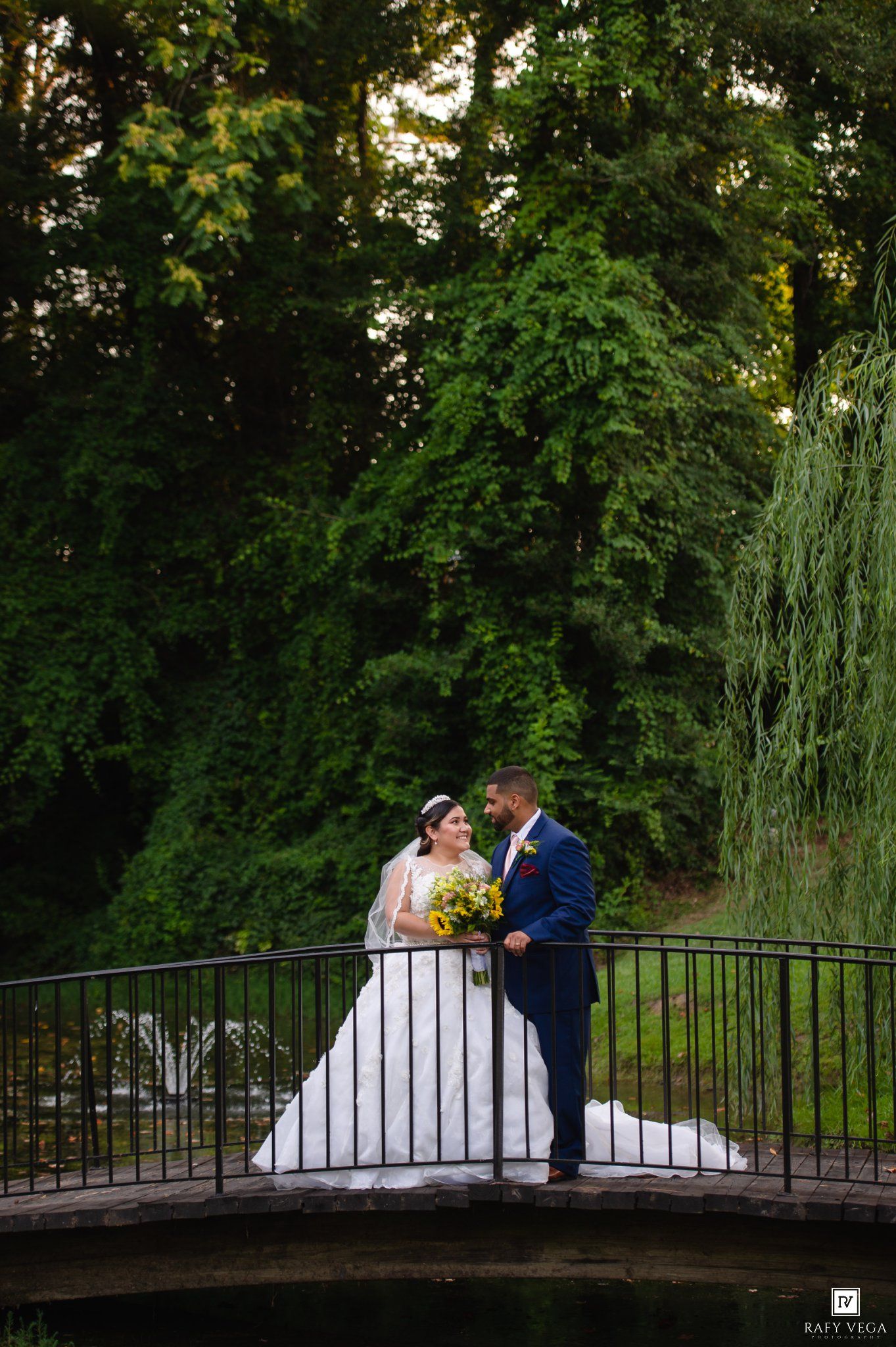 Bride and groom share a romantic moment on a bridge surrounded by lush green trees during their outdoor wedding.