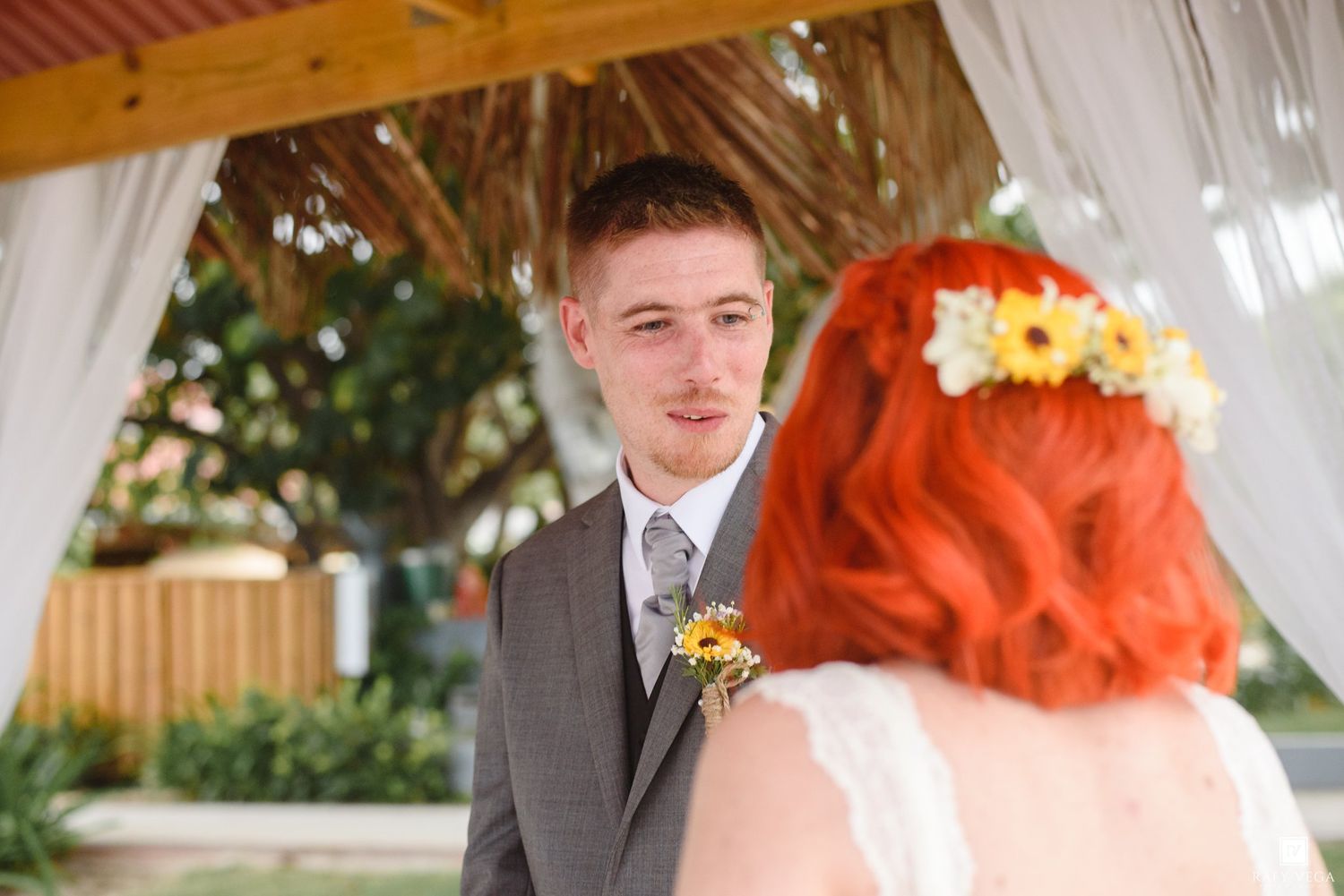 Groom in gray suit exchanges vows with red-haired bride wearing floral crown at outdoor wedding ceremony.