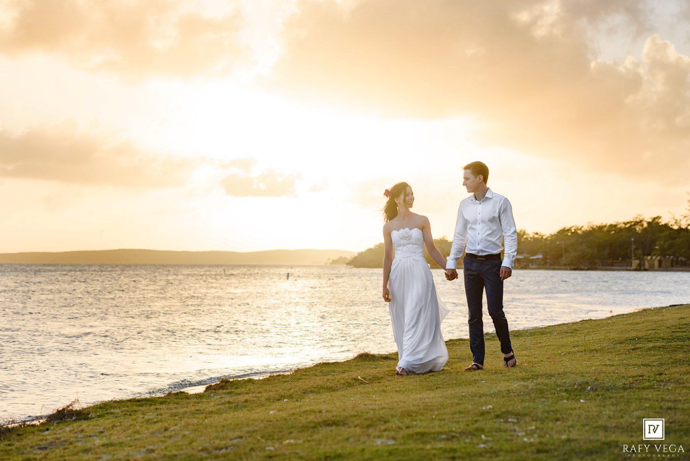 Bride and groom hold hands at golden sunset by the water on their wedding day.