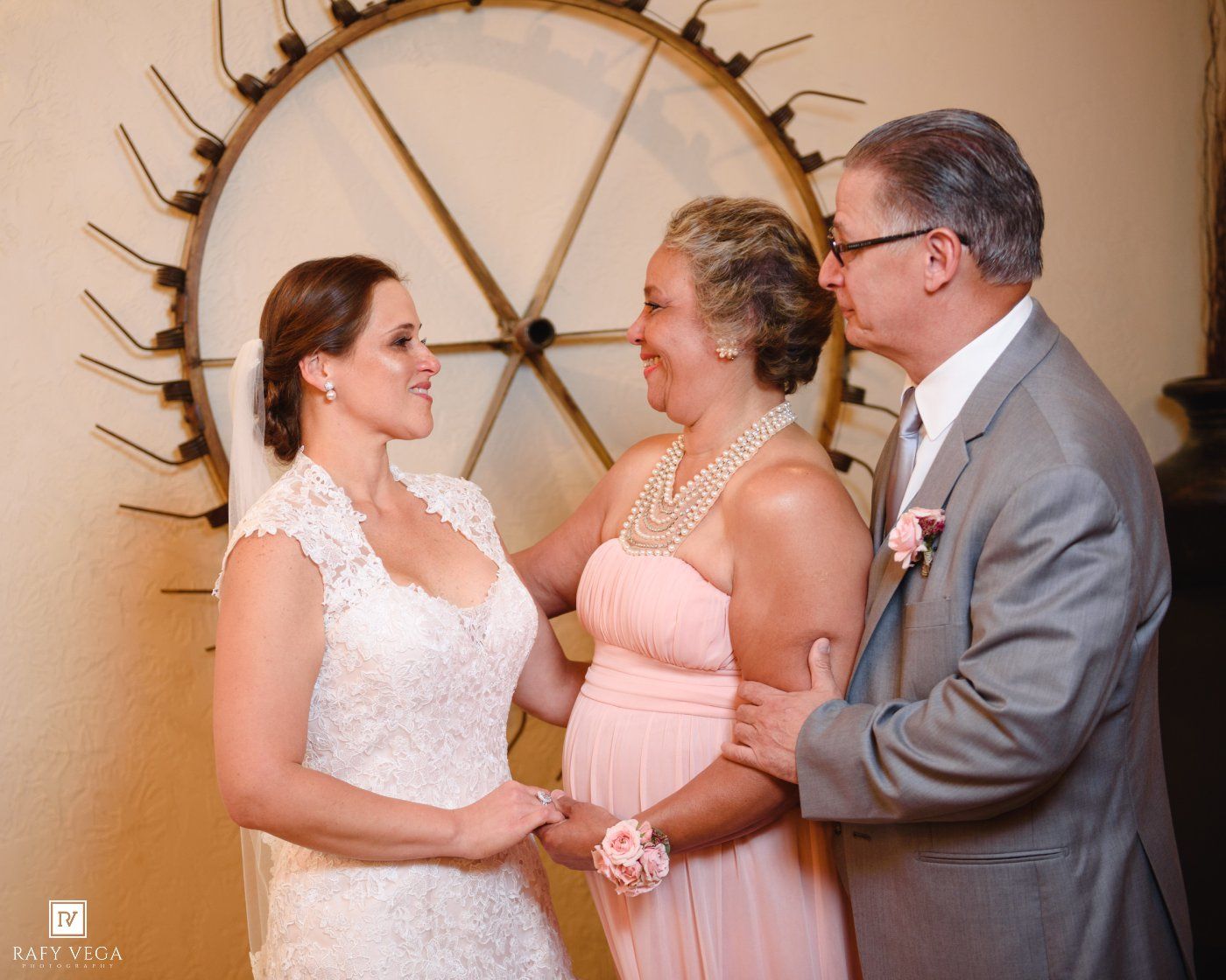 Bride in white lace gown shares a joyful moment with parents in pink dress and gray suit before wedding ceremony.