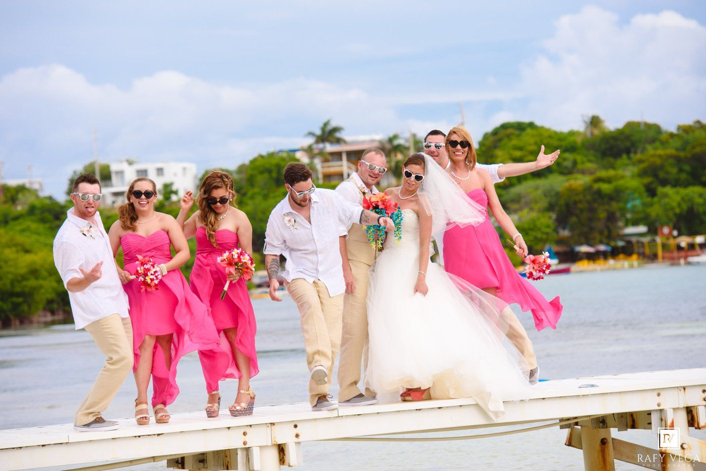 Wedding party walks on a dock in tropical setting, bridesmaids in pink dresses, bride in white gown with arms outstretched.