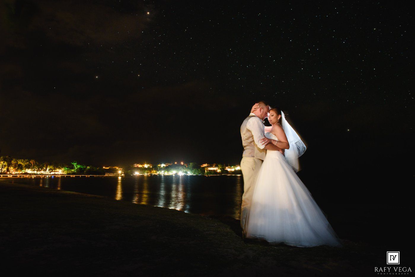 Newlyweds share romantic kiss under starry night sky with ocean lights reflecting behind them.