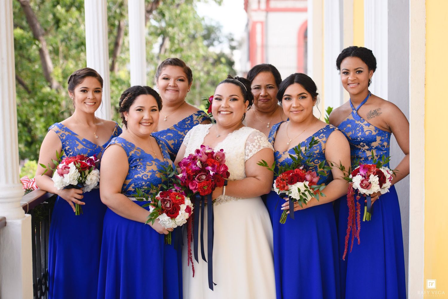 Bride in white gown poses with six bridesmaids in royal blue dresses holding red and white bouquets on a porch.