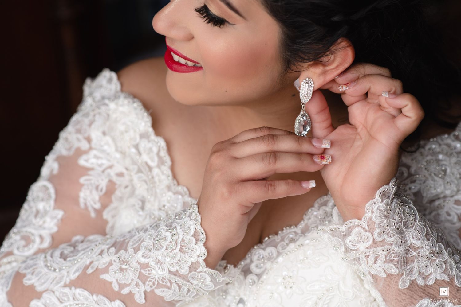 Bride in lace wedding gown adjusting crystal drop earring, smiling with red lips and manicured nails.