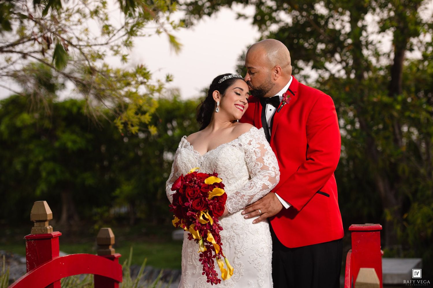 Bride in lace gown holds red bouquet beside groom in red blazer, sharing romantic moment outdoors.