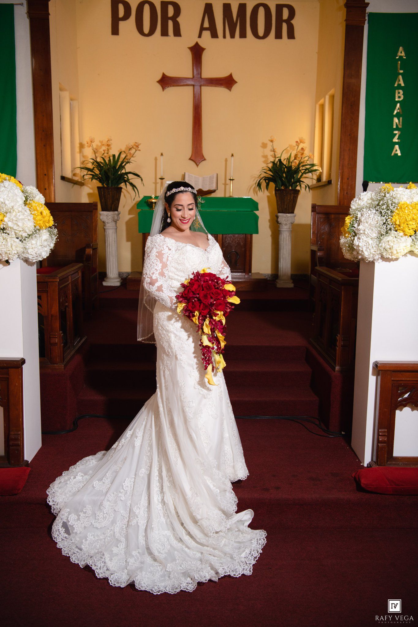 Bride in white lace gown holds red bouquet at church altar with cross and Por Amor sign.