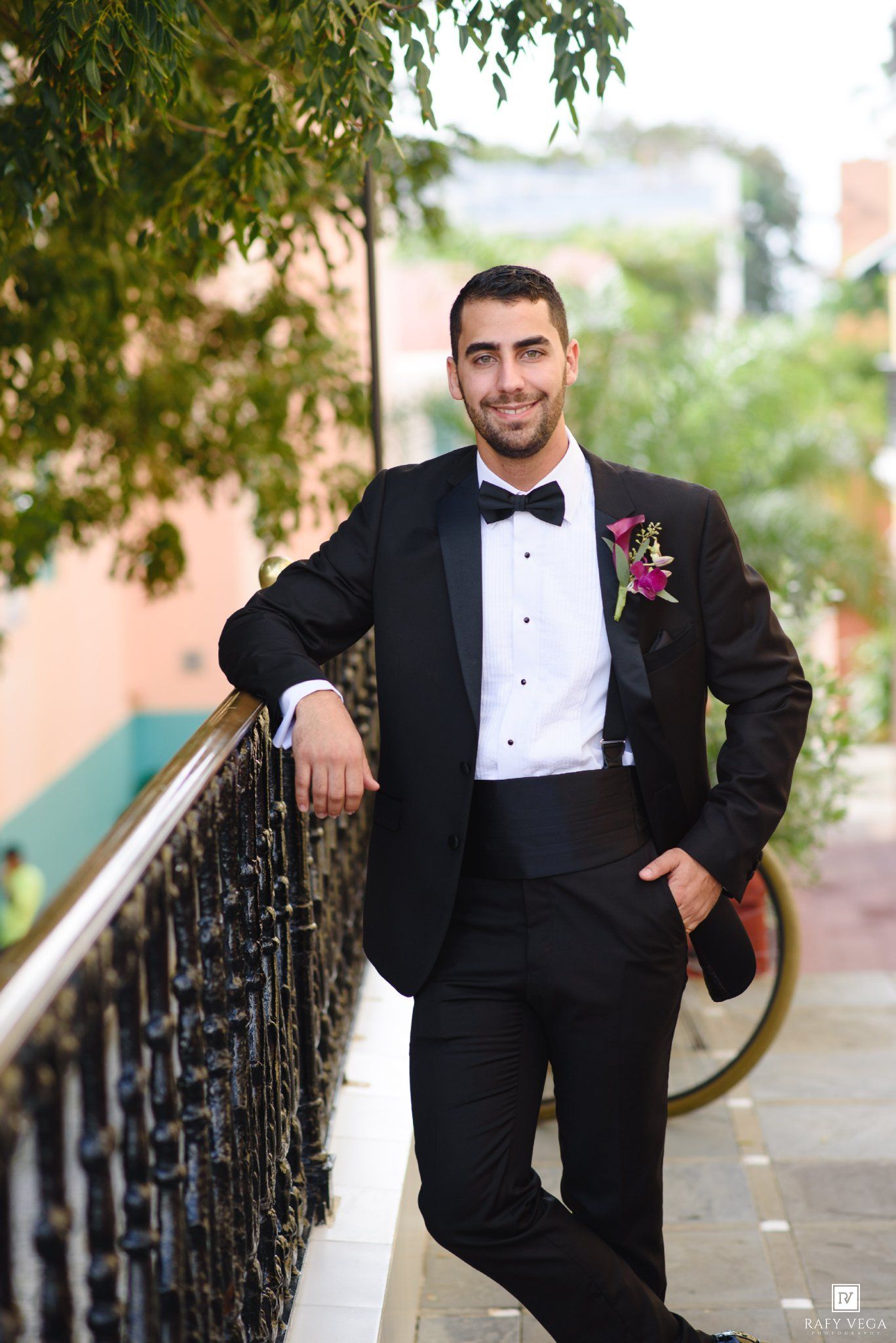 Groom in black tuxedo with bow tie and boutonniere leaning on railing outdoors at wedding venue.