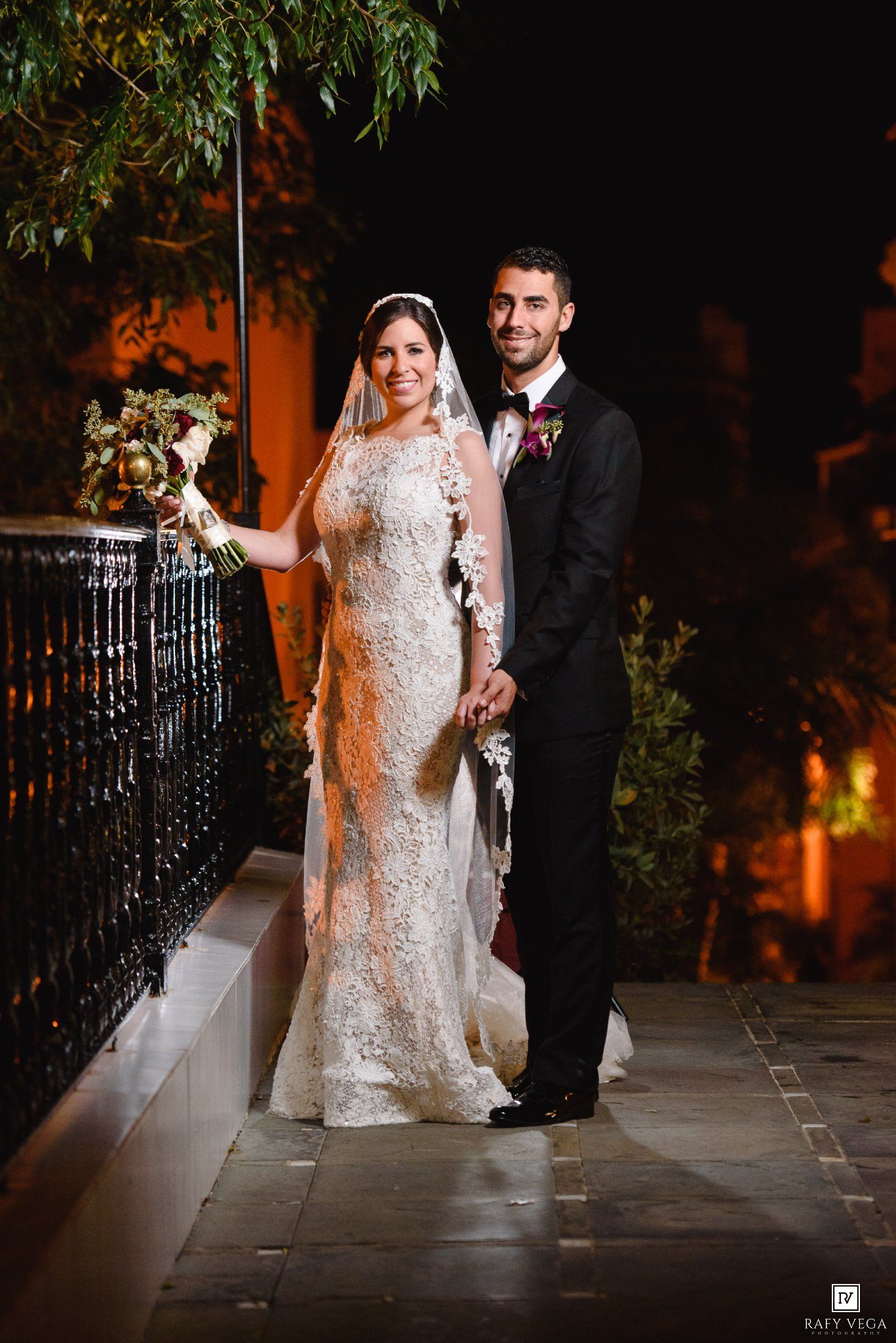 Bride in lace gown with veil and groom in black tuxedo pose together outdoors at night on their wedding day.