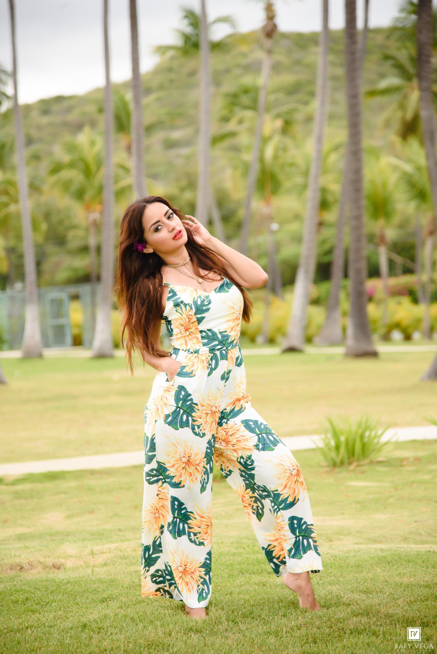 Woman in tropical floral jumpsuit posing on grass with palm trees and green hills in background.