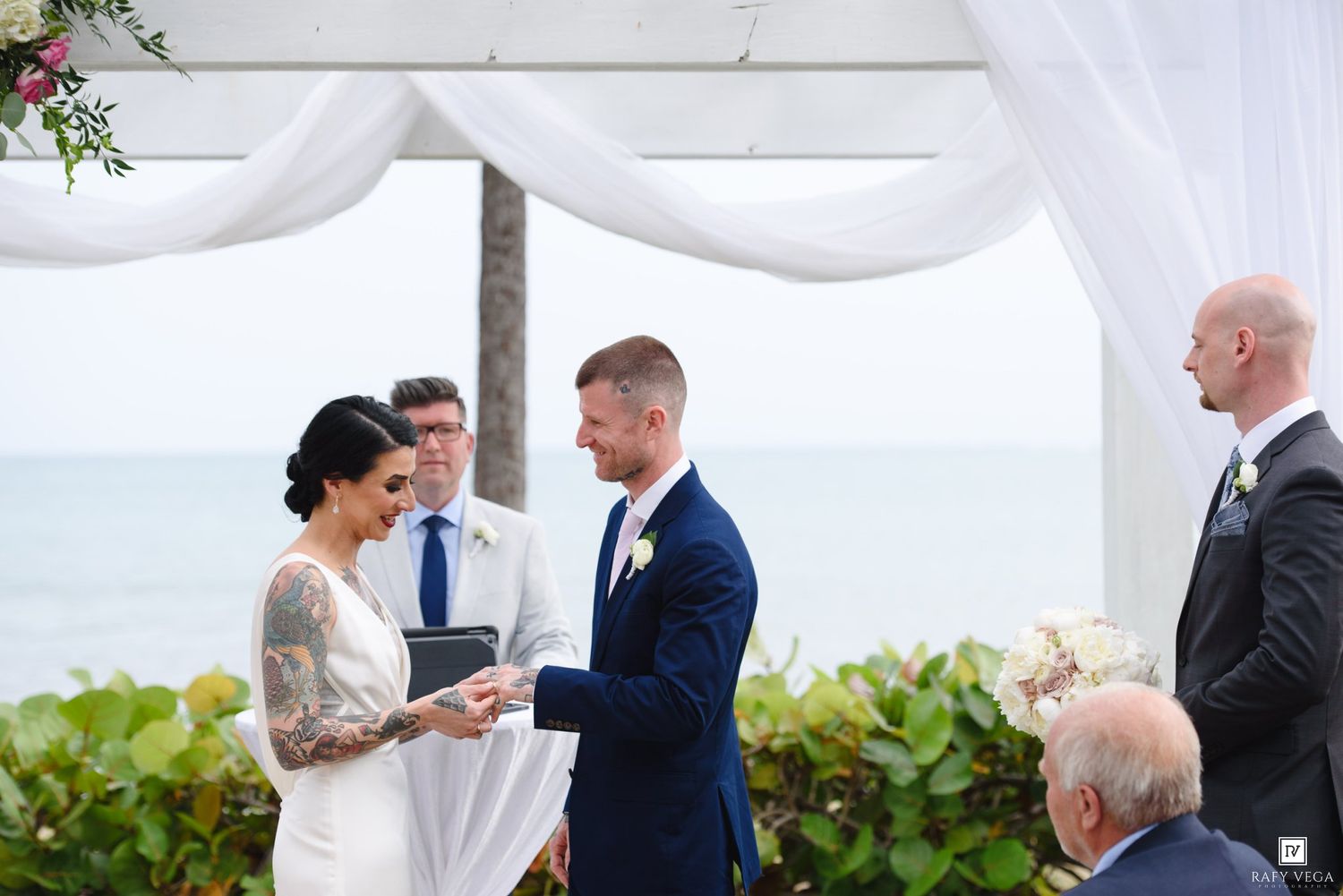 Couple exchanging vows at an outdoor beachfront wedding ceremony under a white draped arch.