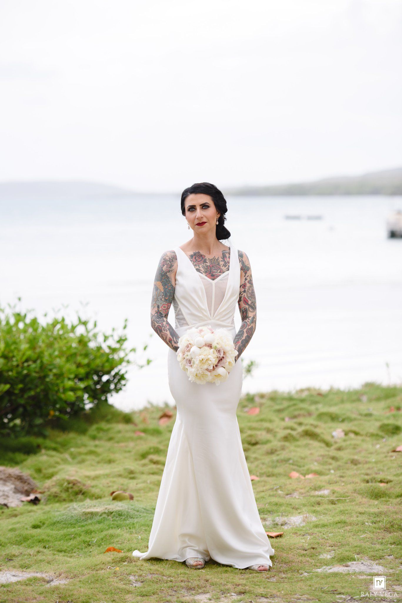 Tattooed bride in a sleeveless white mermaid gown holding a bouquet, standing on a grassy coastal hillside.
