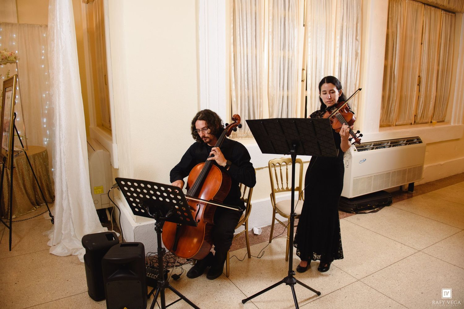 Two musicians in black attire play cello and violin at an elegant indoor event with draped curtains.