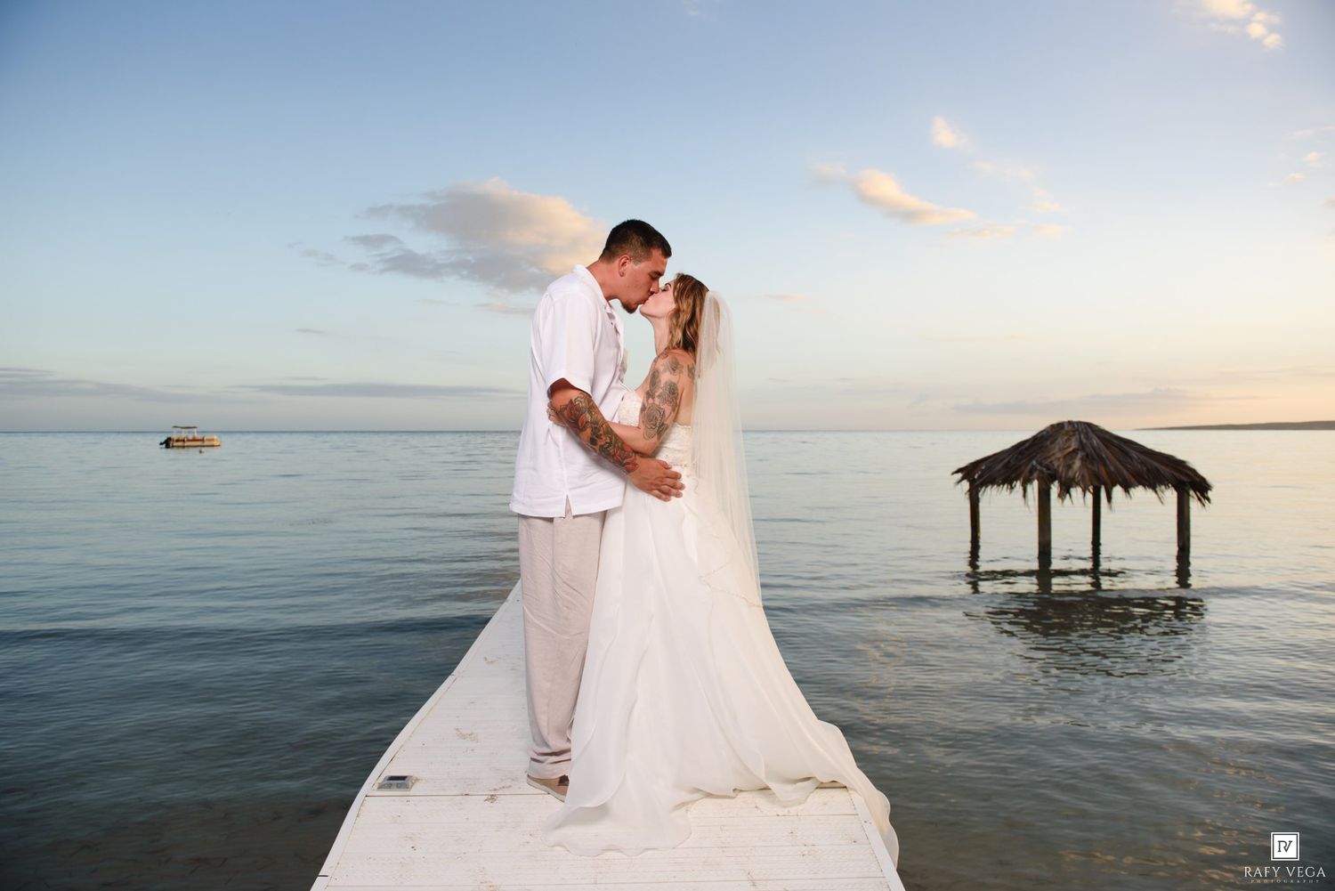 Bride and groom embrace on a pier with a thatched gazebo and calm ocean at sunset.