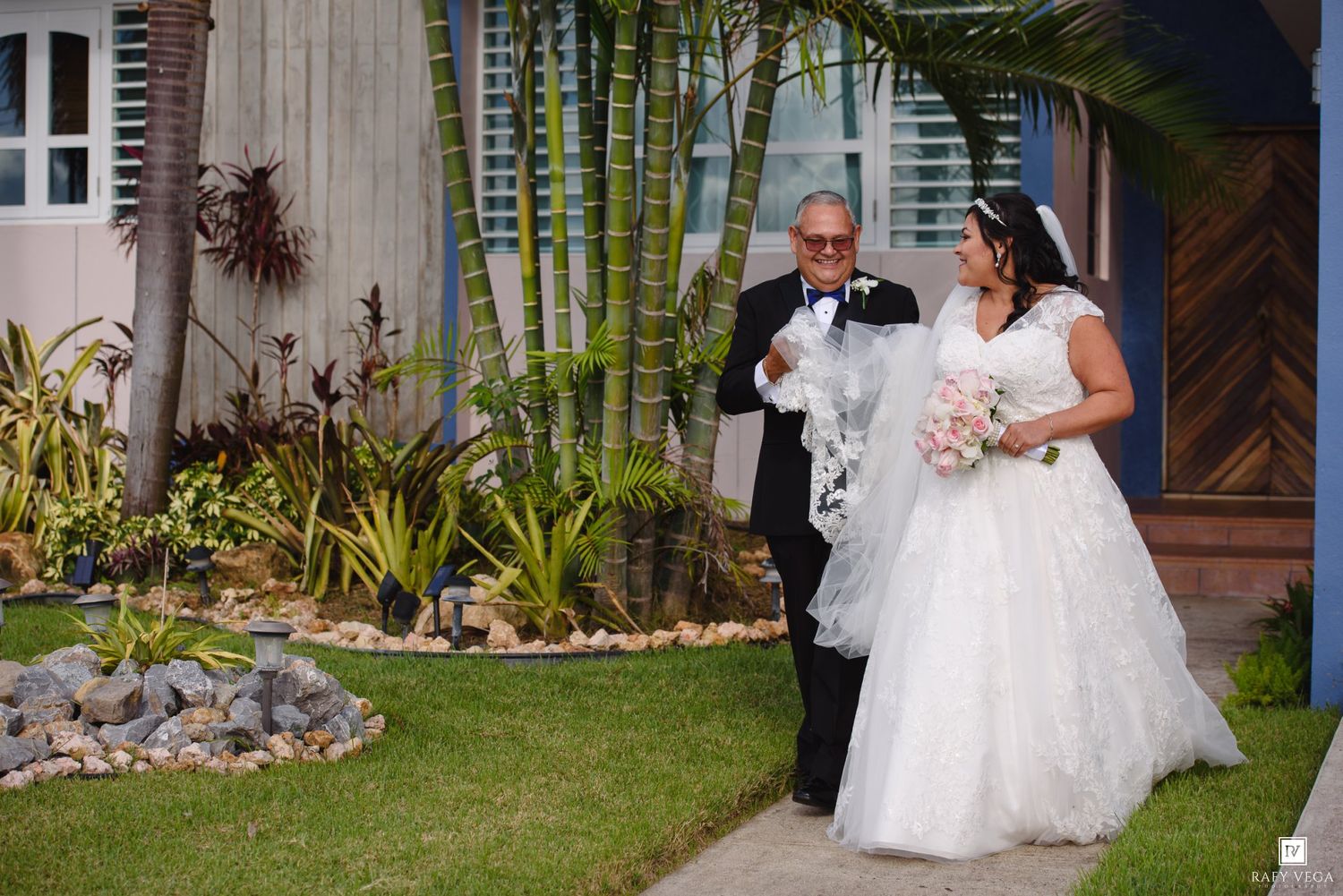 Bride in white ball gown walks with older gentleman past tropical garden toward blue building on wedding day.
